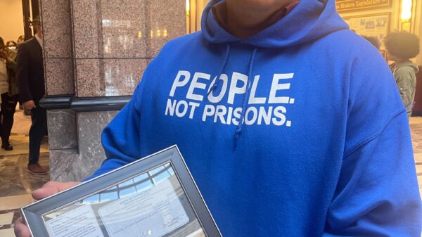 ACLU of CT Smart Justice leader Manuel Sandoval faces the camera, standing and holding a framed collage commemorating his LCSW. he is wearing glasses and a blue people not prisons hoodie