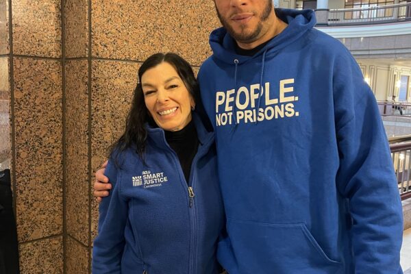 ACLUCT Smart Justice leaders Will and L stand at the capitol, smiling and wearing people not prisons blue hoodies