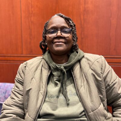 ACLU CT Smart Justice leader Terri Ricks sits, smiling at the camera and wearing a brown jacket and brown hoodie.