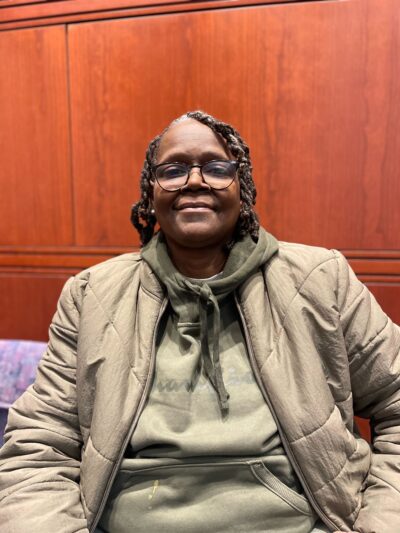 ACLU CT Smart Justice leader Terri Ricks sits, smiling at the camera and wearing a brown jacket and brown hoodie.