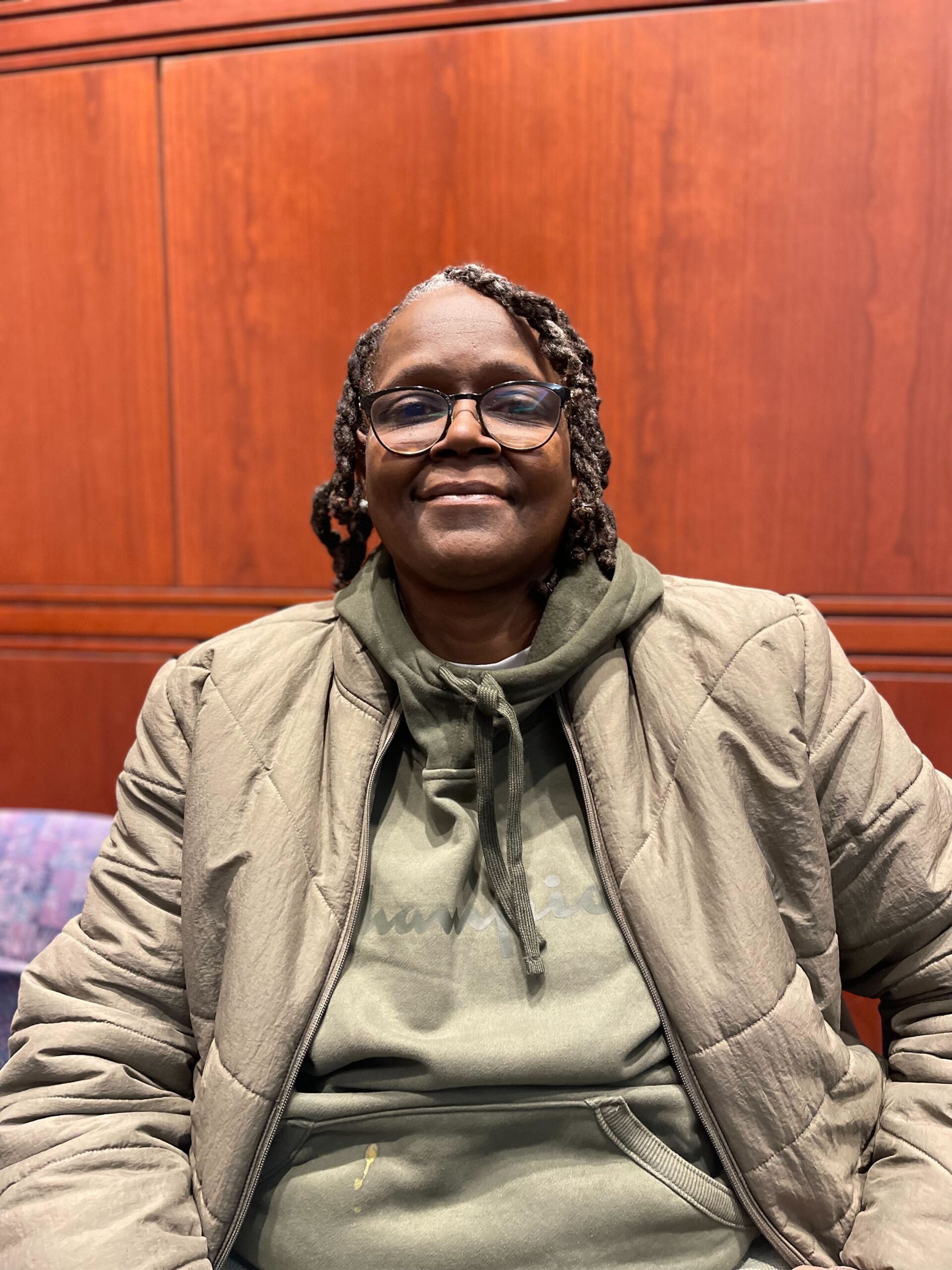 ACLU CT Smart Justice leader Terri Ricks sits, smiling at the camera and wearing a brown jacket and brown hoodie.