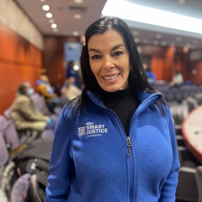 ACLU-CT smart justice leader Lori LeDonne stands, smiling at the camera and wearing a blue zip up sweatshirt. Behind her is a legislative hearing room