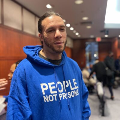 ACLU of CT Smart Justice leader Will Roberts stands, facing the camera, wearing a blue people not prisons hoodie