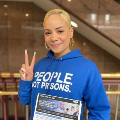 ACLU of CT Smart Justice leader Marquita Reale stands, smiling at the camera and holding up a peace sign. she is wearing a blue people not prisons hoodie