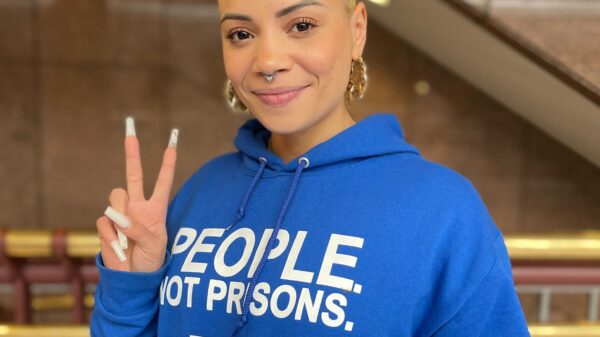 ACLU of CT Smart Justice leader Marquita Reale stands, smiling at the camera and holding up a peace sign. she is wearing a blue people not prisons hoodie