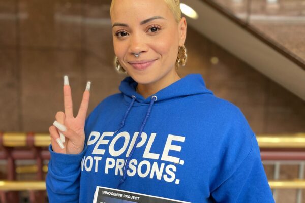 ACLU of CT Smart Justice leader Marquita Reale stands, smiling at the camera and holding up a peace sign. she is wearing a blue people not prisons hoodie