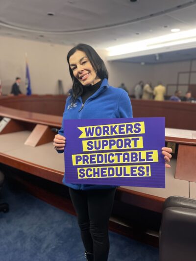 ACLUCT Smart Justice leader Lori LeDonne stands, smiling and holding a "workers support predictable schedules" sign. She is wearing a blue people not prisons zip up sweatshirt