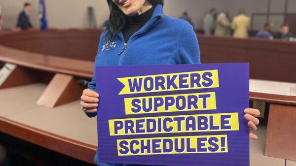ACLUCT Smart Justice leader Lori LeDonne stands, smiling and holding a "workers support predictable schedules" sign. She is wearing a blue people not prisons zip up sweatshirt