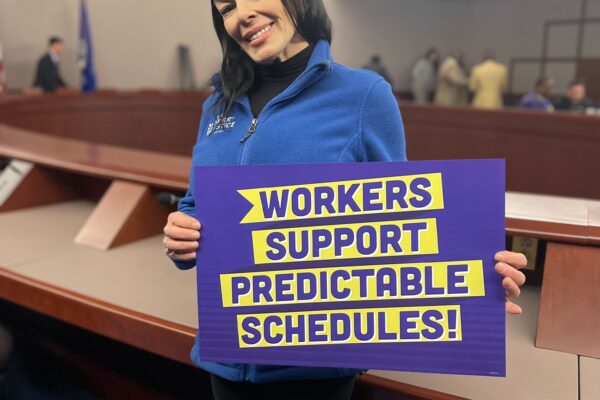 ACLUCT Smart Justice leader Lori LeDonne stands, smiling and holding a "workers support predictable schedules" sign. She is wearing a blue people not prisons zip up sweatshirt
