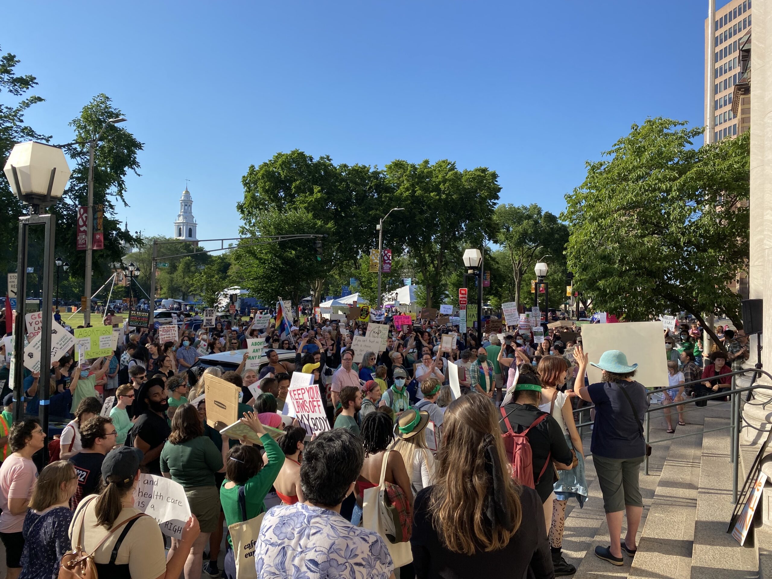 Protesters gather in New Haven after the US Supreme Court gutted Roe v Wade in the Dobbs decision. The sky is blue and clear, people are holding signs and trees are visible in the background