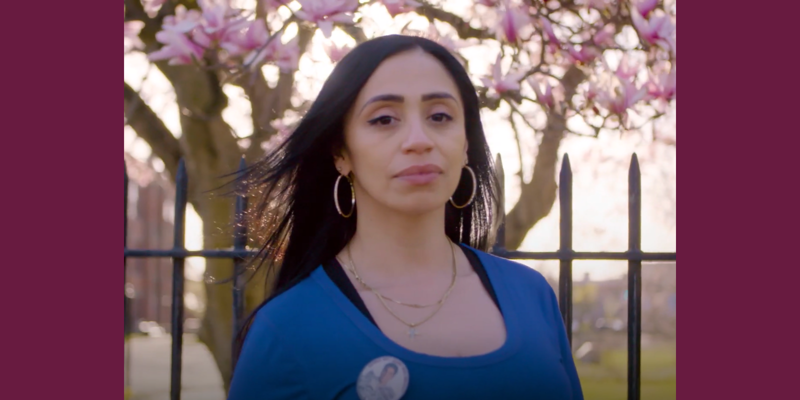 Natasha Tosado, a woman with long brown hair, faces the camera. She looks powerful and serious, and she is wearing a blue shirt and a pin with a photo of her son, Jayson, on it. Behind her is a fence and a blooming magnolia tree.