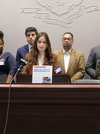 Jess Zaccagnino, a woman with long brown hair, stands behind a podium. In front of her is a sign that says "CT voting rights act." Behind her is a group of people, watching as she speaks