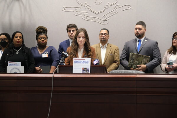 Jess Zaccagnino, a woman with long brown hair, stands behind a podium. In front of her is a sign that says "CT voting rights act." Behind her is a group of people, watching as she speaks