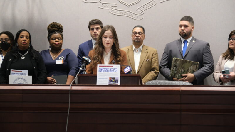 Jess Zaccagnino, a woman with long brown hair, stands behind a podium. In front of her is a sign that says "CT voting rights act." Behind her is a group of people, watching as she speaks