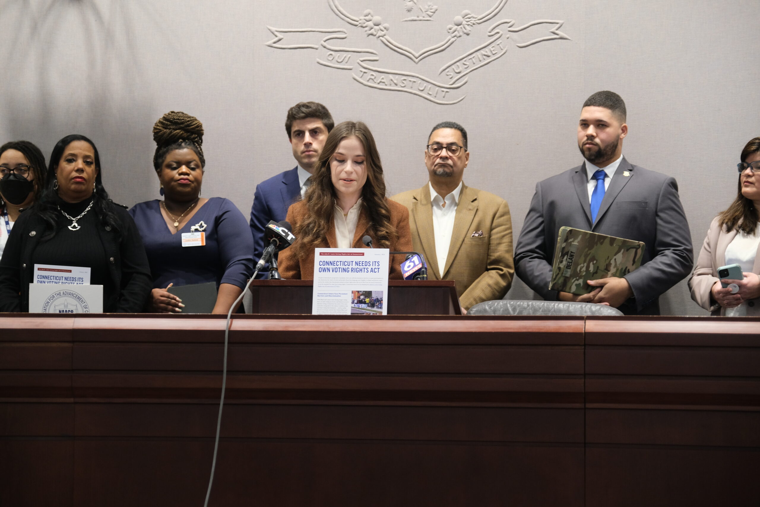 Jess Zaccagnino, a woman with long brown hair, stands behind a podium. In front of her is a sign that says "CT voting rights act." Behind her is a group of people, watching as she speaks