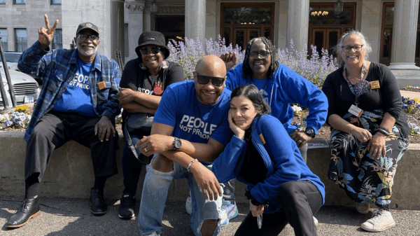 Six ACLU-CT Smart Justice leaders and friends sit and crouch in front of the CT Capitol building. They are smiling, wearing blue "people not prisons" shirts, and the sun is shining.