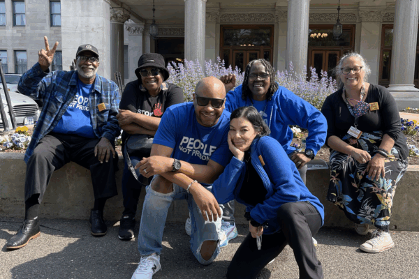 Six ACLU-CT Smart Justice leaders and friends sit and crouch in front of the CT Capitol building. They are smiling, wearing blue "people not prisons" shirts, and the sun is shining.