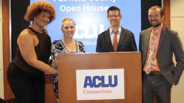 Behind a wooden podium with the ACLU-CT's logo, stands a Black-Taino women with a light brown afro on the left, a white woman with blonde hair to her right, a white man with dark hair and glasses to her right, and a white man with brown hair on the right.
