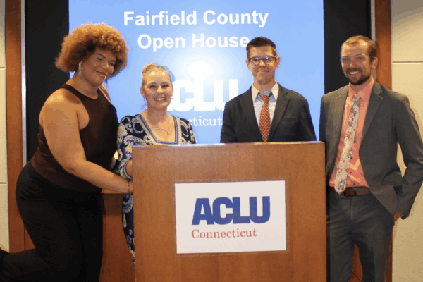 Behind a wooden podium with the ACLU-CT's logo, stands a Black-Taino women with a light brown afro on the left, a white woman with blonde hair to her right, a white man with dark hair and glasses to her right, and a white man with brown hair on the right.