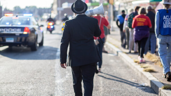 In the road, a person in a dark suit with a hat appears to be joining a protest against police with a megaphone. A police car is to their left.