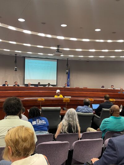 This photo captures a legislative forum about the fake traffic ticket scandal from the back of the room. Several people's backs are towards the camera with legislators in the background of the photo.