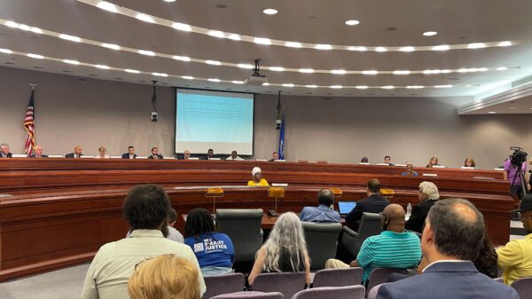 This photo captures a legislative forum about the fake traffic ticket scandal from the back of the room. Several people's backs are towards the camera with legislators in the background of the photo.