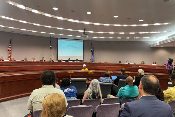 This photo captures a legislative forum about the fake traffic ticket scandal from the back of the room. Several people's backs are towards the camera with legislators in the background of the photo.
