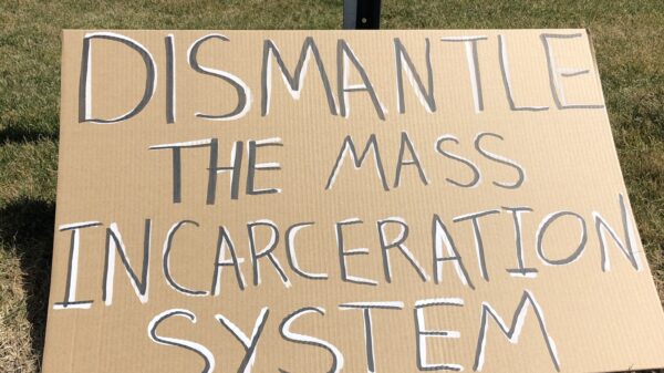A cardboard sign sits on the grass in front of the CT state capitol building. In black letters with white background, the sign says: "dismantle the mass incarceration system"