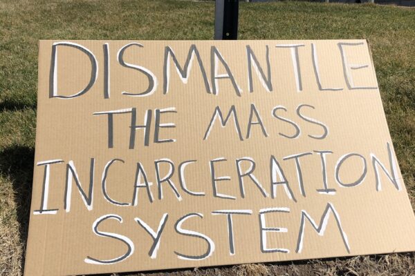A cardboard sign sits on the grass in front of the CT state capitol building. In black letters with white background, the sign says: "dismantle the mass incarceration system"