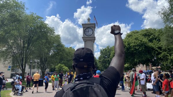 A crowd of people is gathered around the clock tower in Waterbury, Connecticut. The sky is blue and it is sunny. A Black man is in the foreground, back to the camera, with fist raised. He is wearing a backpack.