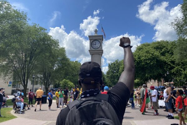 A crowd of people is gathered around the clock tower in Waterbury, Connecticut. The sky is blue and it is sunny. A Black man is in the foreground, back to the camera, with fist raised. He is wearing a backpack.