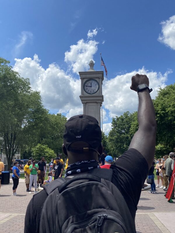 A crowd of people is gathered around the clock tower in Waterbury, Connecticut. The sky is blue and it is sunny. A Black man is in the foreground, back to the camera, with fist raised. He is wearing a backpack.