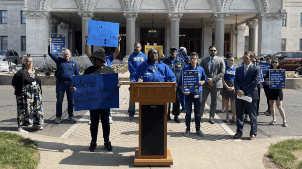 Smart Justice leaders are gathered in front of the Capitol to speak about prosecutorial accountability and one leader, in the center, stands at the podium as they speak.