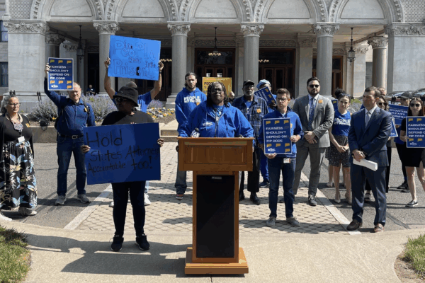 Smart Justice leaders are gathered in front of the Capitol to speak about prosecutorial accountability and one leader, in the center, stands at the podium as they speak.