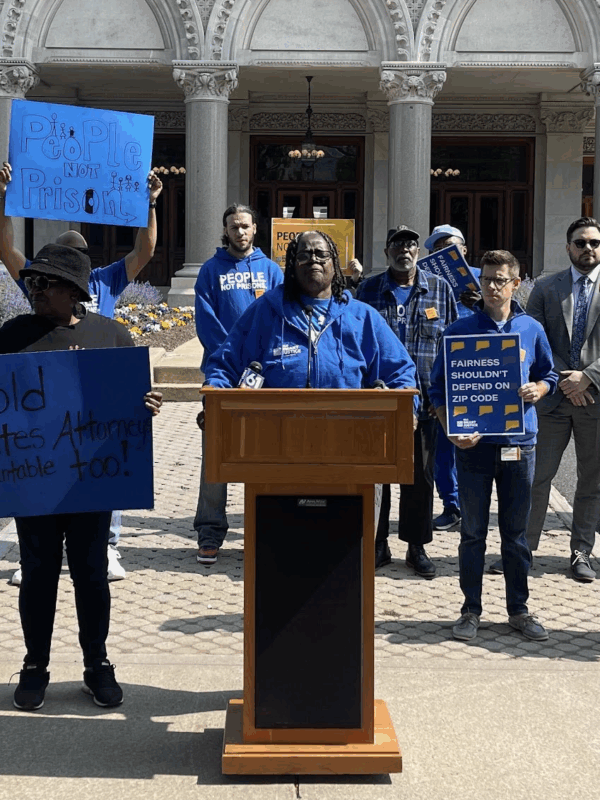 Smart Justice leaders are gathered in front of the Capitol to speak about prosecutorial accountability and one leader, in the center, stands at the podium as they speak.