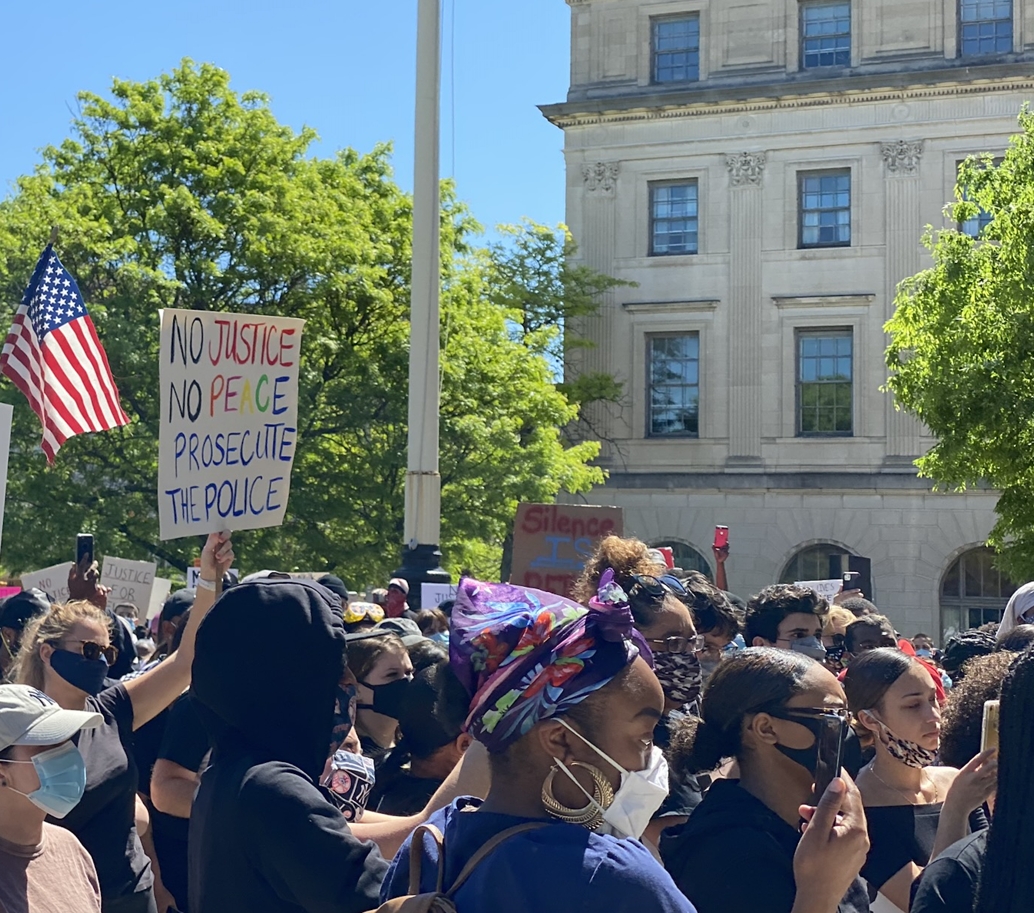 A crowd of protesters walk past the camera. The focus is on a sign that says "No Justice, No Peace, Prosecute the Police."