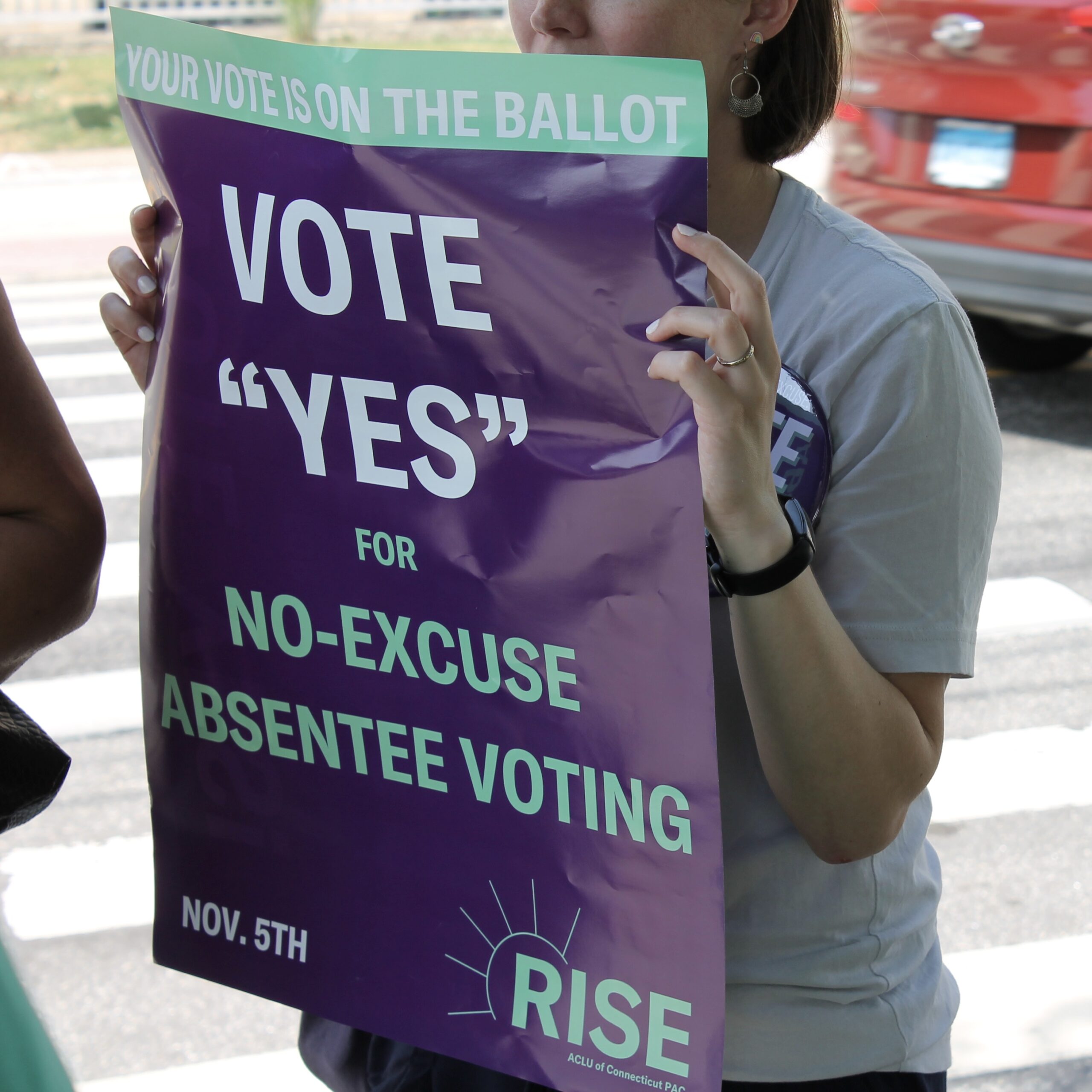 A woman, who is mostly out of frame, is holding a purple sign that says "VOTE "YES" FOR NO-EXCUSE ABSENTEE VOTING" in white and teal text.