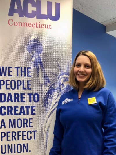 ACLUCT smart justice leader Tracie Bernardi stands facing the camera and smiling. She is wearing a blue Smart Justice zip-up and a yellow People Not Prisons pin. To her left is an ACLUCT banner that says we the people dare to create a more perfect union
