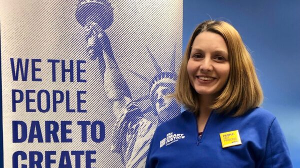 ACLUCT smart justice leader Tracie Bernardi stands facing the camera and smiling. She is wearing a blue Smart Justice zip-up and a yellow People Not Prisons pin. To her left is an ACLUCT banner that says we the people dare to create a more perfect union