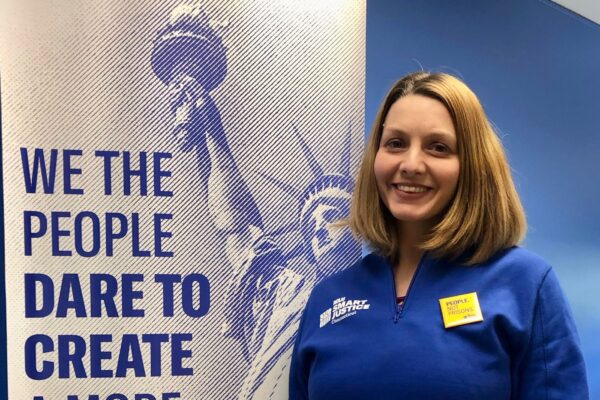 ACLUCT smart justice leader Tracie Bernardi stands facing the camera and smiling. She is wearing a blue Smart Justice zip-up and a yellow People Not Prisons pin. To her left is an ACLUCT banner that says we the people dare to create a more perfect union