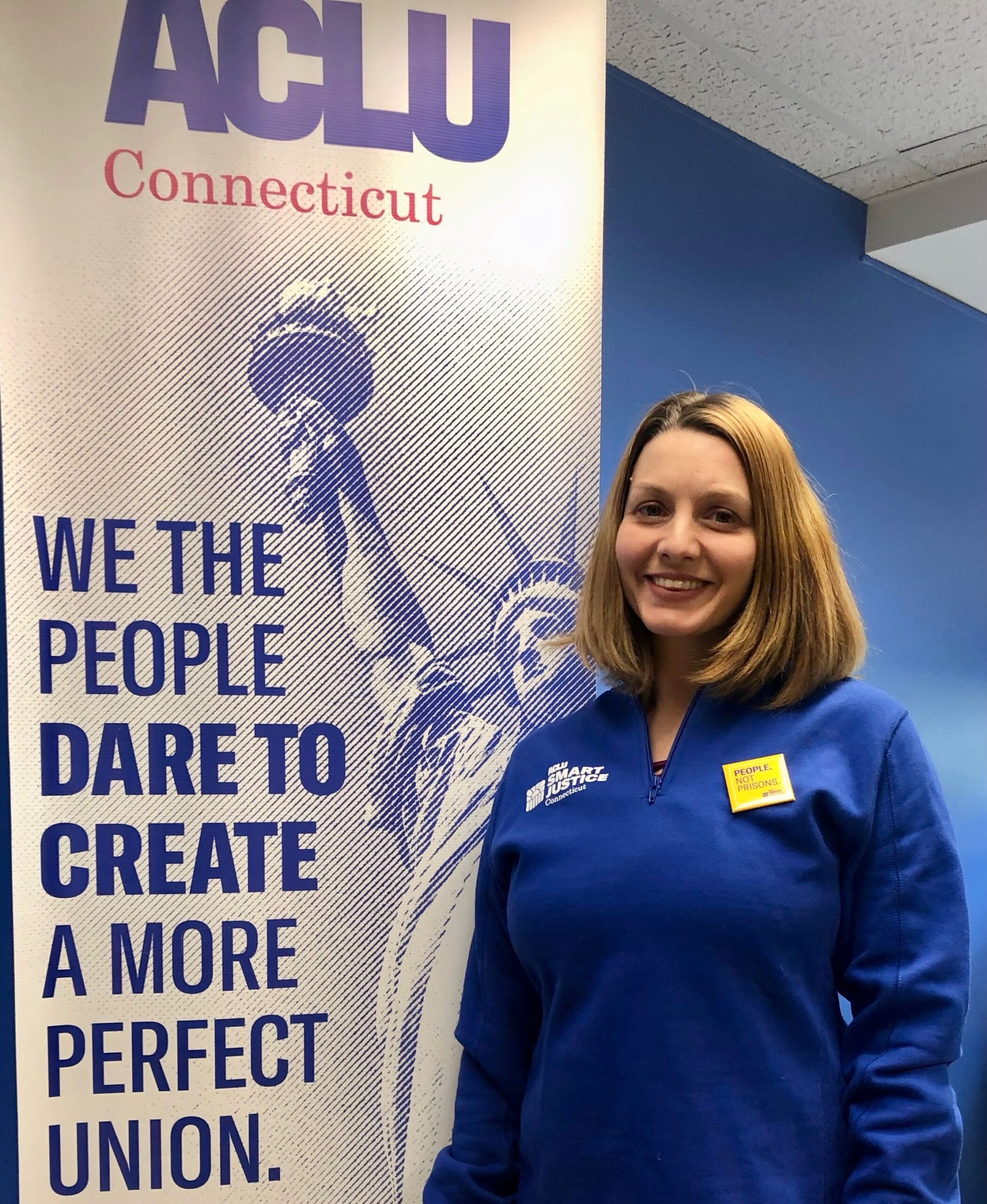 ACLUCT smart justice leader Tracie Bernardi stands facing the camera and smiling. She is wearing a blue Smart Justice zip-up and a yellow People Not Prisons pin. To her left is an ACLUCT banner that says we the people dare to create a more perfect union
