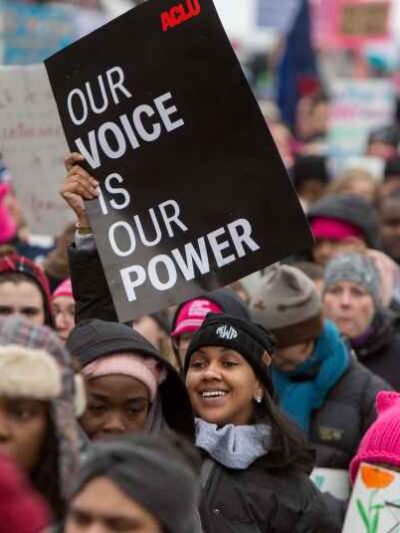 There is a crowd of folks rallying for reproductive justice. The focus is on one femme-presenting person with a beanie and a sign up in the air that is a black poster that says "Our Voice is Our Power."