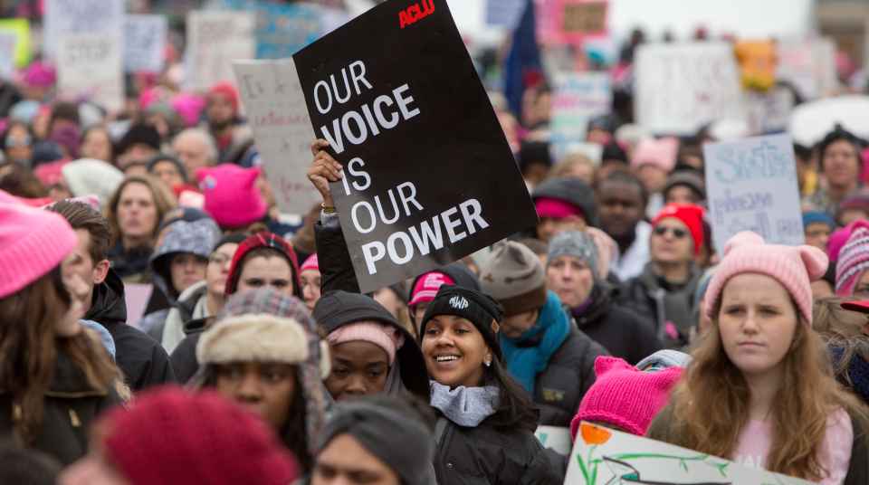 There is a crowd of folks rallying for reproductive justice. The focus is on one femme-presenting person with a beanie and a sign up in the air that is a black poster that says "Our Voice is Our Power."