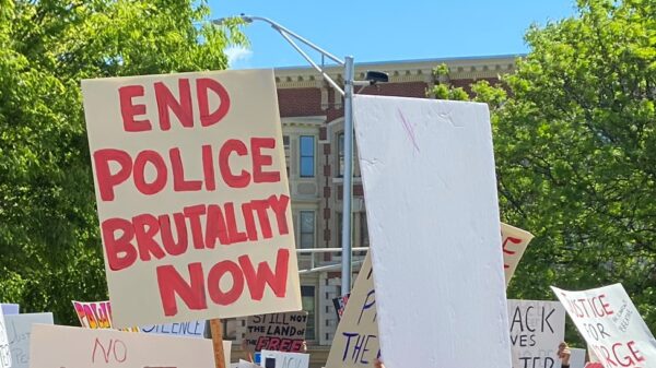 A white sign with red writing, surrounded by other protest signs and against a backdrop of trees. The sign with red writing says: END POLICE BRUTALITY NOW