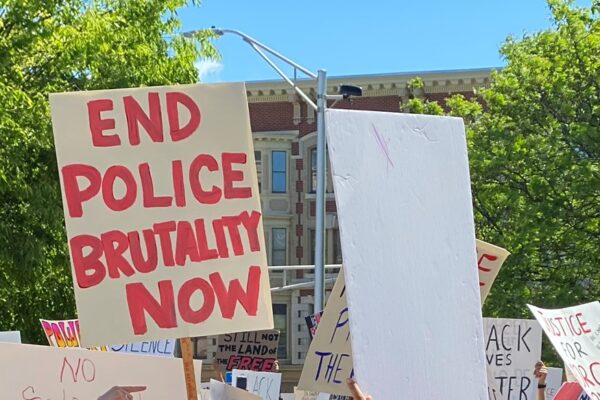A white sign with red writing, surrounded by other protest signs and against a backdrop of trees. The sign with red writing says: END POLICE BRUTALITY NOW