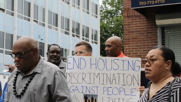 A crowd stands in front of a rental management building with signs to advocate for equitable housing. Gus, our campaign manager, is in the center in focus with a sign that says "end housing discrimination against people with a record of conviction."