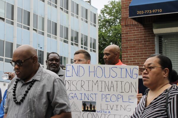 A crowd stands in front of a rental management building with signs to advocate for equitable housing. Gus, our campaign manager, is in the center in focus with a sign that says "end housing discrimination against people with a record of conviction."