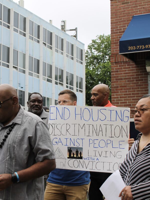 A crowd stands in front of a rental management building with signs to advocate for equitable housing. Gus, our campaign manager, is in the center in focus with a sign that says "end housing discrimination against people with a record of conviction."