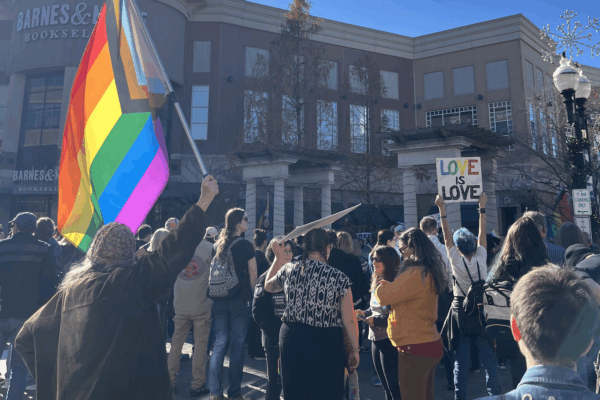 In front of a Barnes & Nobles, a crowd stands in front of a stage. Several people are holding signs, LGBTQIA+ flags, etc.