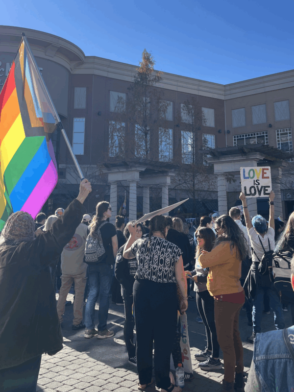 In front of a Barnes & Nobles, a crowd stands in front of a stage. Several people are holding signs, LGBTQIA+ flags, etc.